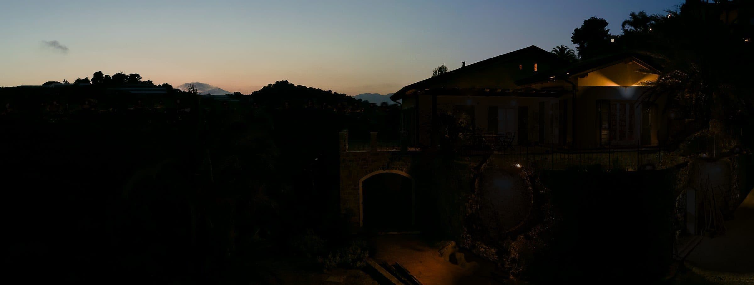 A hillside house at dusk with warm lights in its windows, distant village lights along the ridge